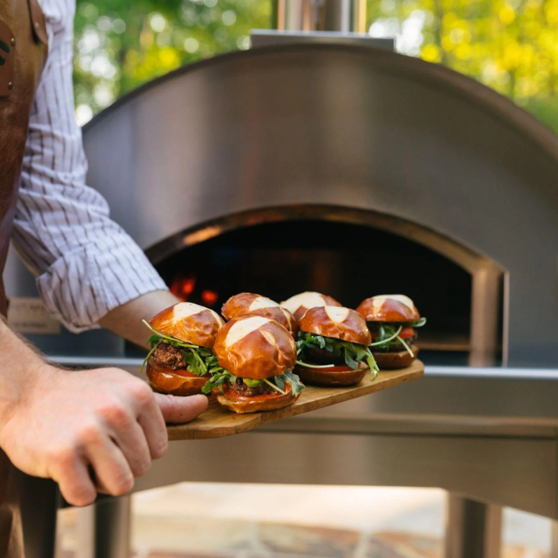 Person holding a wooden board with sandwiches in front of an outdoor pizza oven. Fontana Forni Marinara Pizza Oven Dual Fuel Countertop